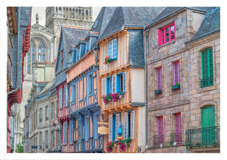 Colorful historic buildings line a quaint street in an old French town, with flowers and cathedral in the background.