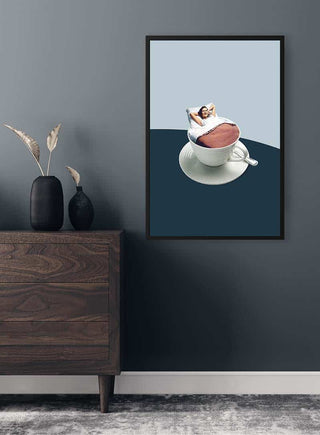 Framed art print of a woman relaxing in a coffee cup displayed on a dark wall above a wooden dresser.