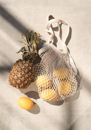 A pineapple and several yellow summer fruits in a mesh bag on a light surface.