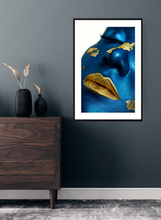 Framed poster of a face painted blue with metallic golden lips and gold accents, hanging on a dark wall.