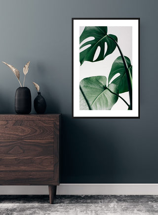 Monstera leaves poster in a black frame displayed on a dark wall above a wooden dresser.