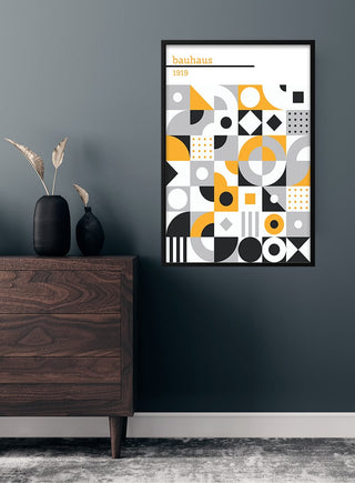 Bauhaus 1919 black and white poster with geometric shapes, framed on a dark wall above a wooden dresser.
