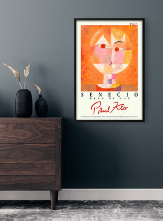 Framed Paul Klee "Head of Man" poster on a dark wall above a wooden dresser with decorative vases.