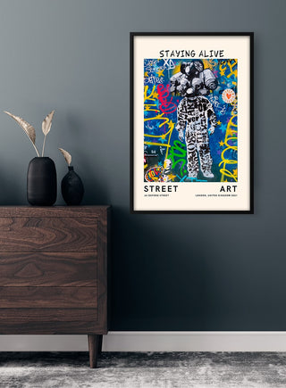 Framed "Staying Alive" street art poster with vibrant graffiti on a dark gray wall above a wooden dresser.