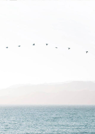Minimalist artwork of birds flying in a straight line over calm water with soft mountains in the background.
