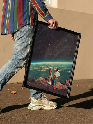 Person holding a framed "Love - Frank Moth" artwork featuring a couple gazing at Earth from space.