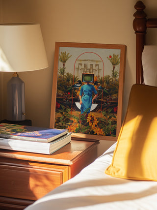 Framed "Her Asymmetries" artwork by Frank Moth on a bedside table beside books and a lamp.