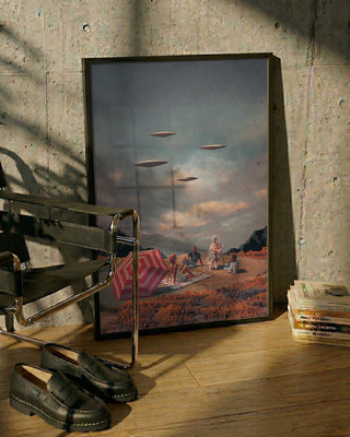 Framed "Pretend They Never Came" by Frank Moth, featuring UFOs over a picnic scene, leaning against a wall.