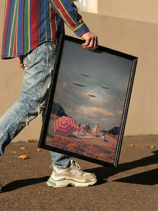 Person holding framed "Pretend They Never Came" artwork by Frank Moth, with UFOs over a picnic scene.
