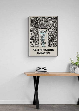 Framed "Humanism" print by Keith Haring displayed above a wooden desk with plants and books.