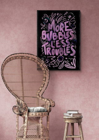 Framed "More Bubbles Less Troubles" Baroo Bloom print on pink wall above wicker chair and small stool with books.