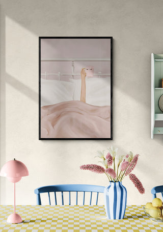 Framed art of a hand holding a coffee cup in bed, styled above a dining table with flowers and pastel decor.