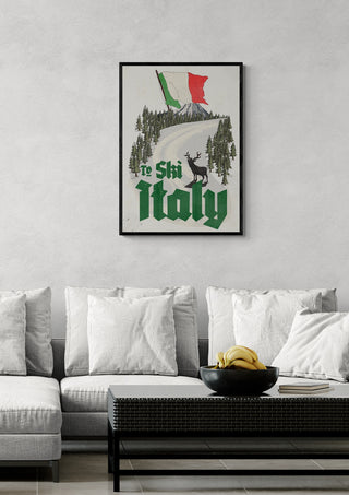 Vintage postcard print with "To Ski Italy," trees, a deer, and flags, displayed above a modern sofa.