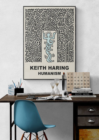 Framed "Humanism" poster by Keith Haring hanging above a modern desk with books and decor items.